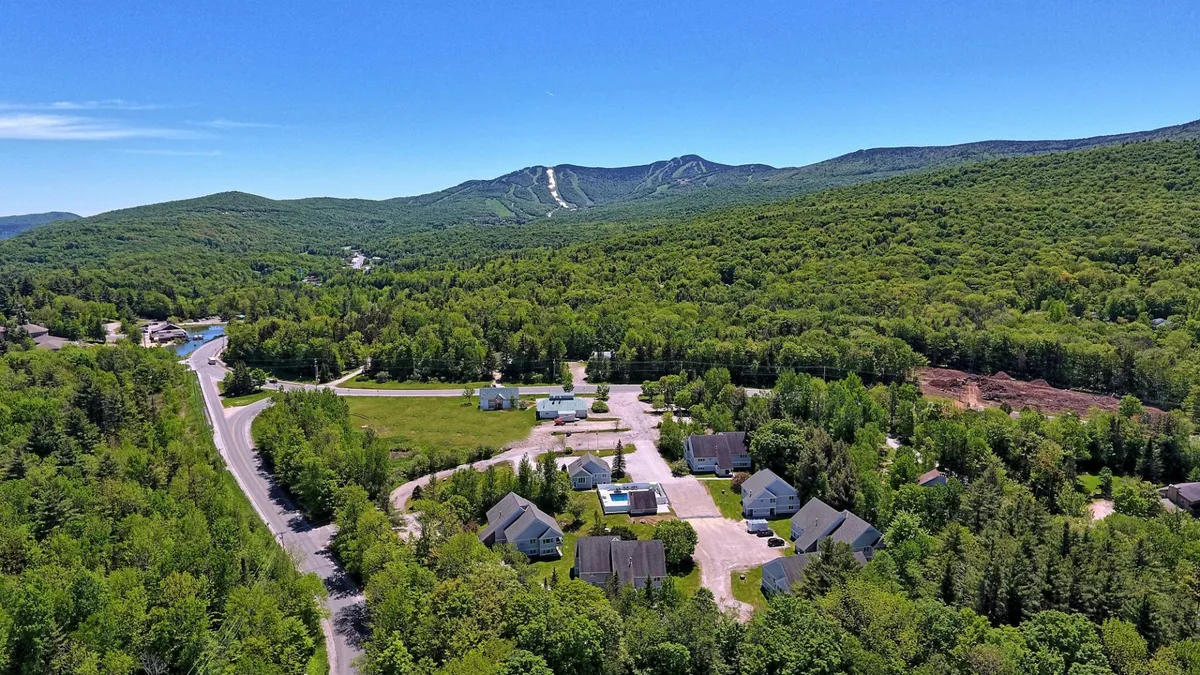 This exterior image features a scenic view of mountains in the background, residential buildings in the midground, and roadways winding through the area. The landscape is tranquil, highlighting the proximity of the complex to nature and the mountain slopes.
