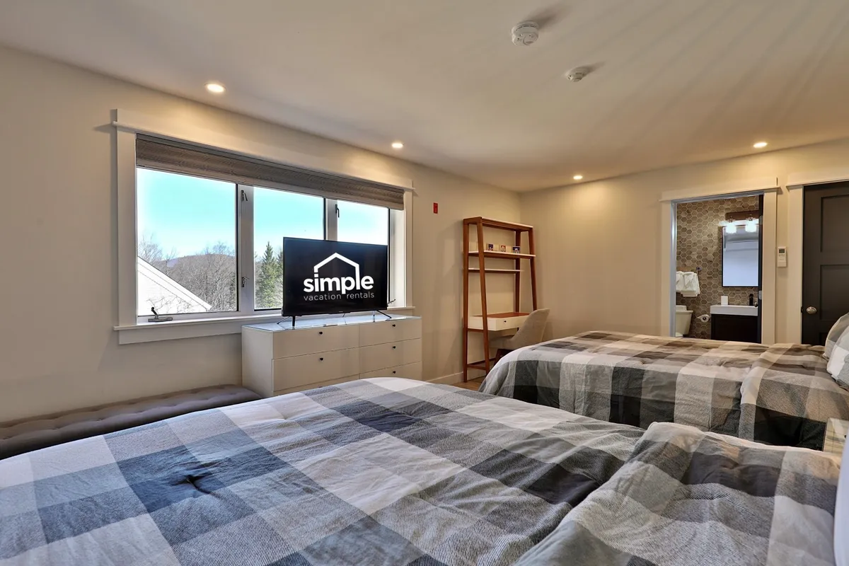 A spacious bedroom featuring two plush queen beds with checkered blankets, a television displaying the suite's logo, and a window offering a glimpse of the surrounding natural landscape. The arrangement suggests a setting perfect for relaxing together, watching TV as a group, or planning excursions.