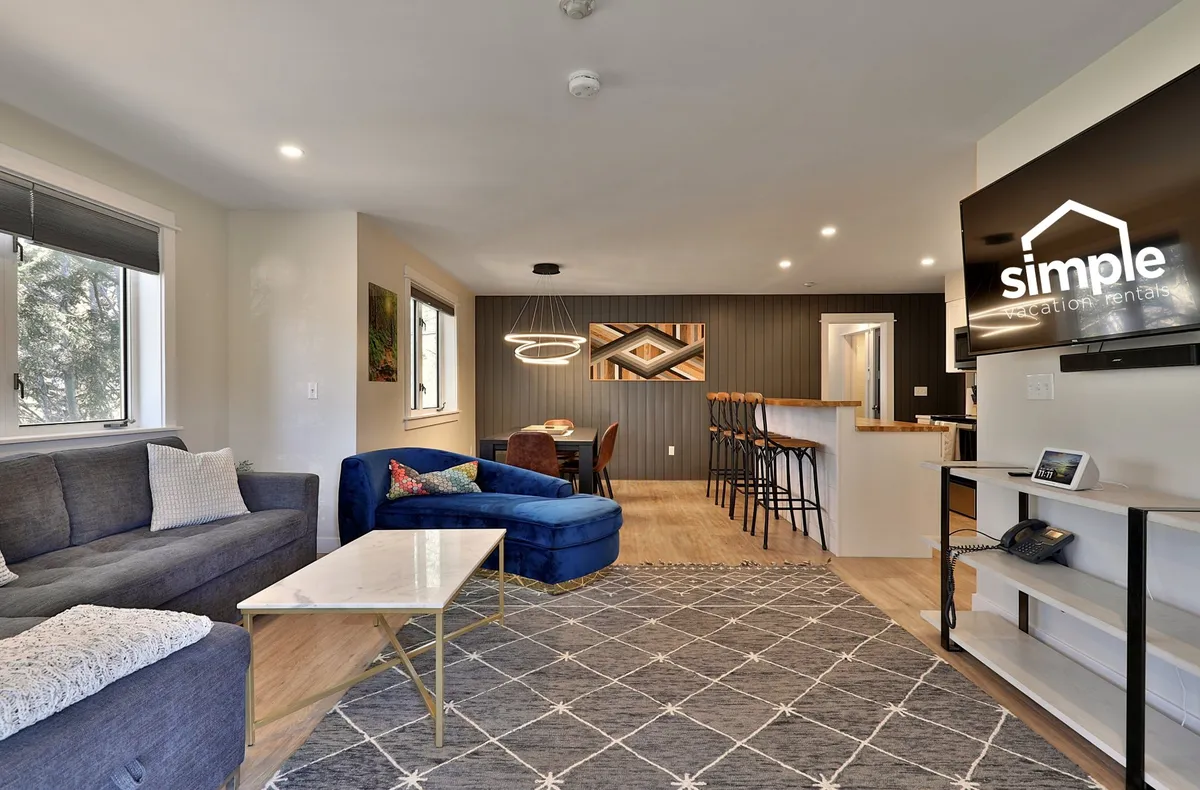 A bright, open-concept living area boasting a comfortable sofa and blue chair centered around a coffee table, with bar stools at the kitchen counter and a TV on the wall. The space is designed for both relaxation and socializing, with ample seating and a modern, airy vibe.