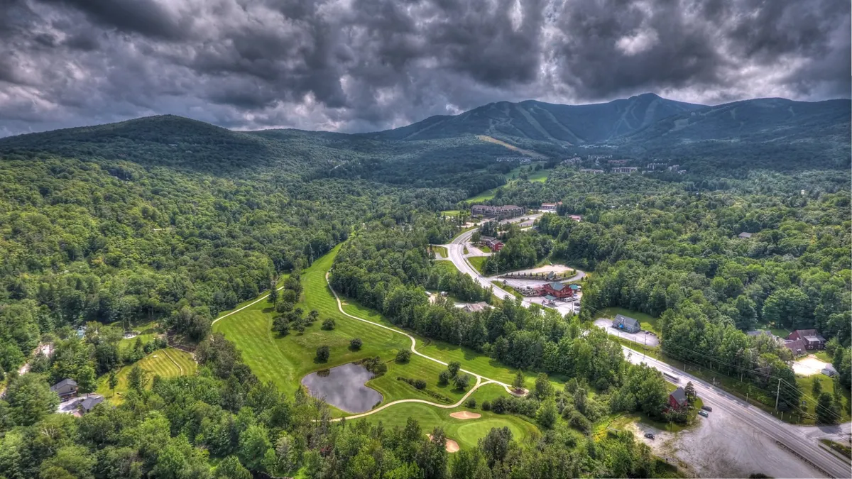 A wide scenic view featuring green mountains, a lush golf course, a tranquil pond, and a winding road. The image captures the essence of outdoor summer activities with a clear view of both natural landscapes and recreational amenities.
