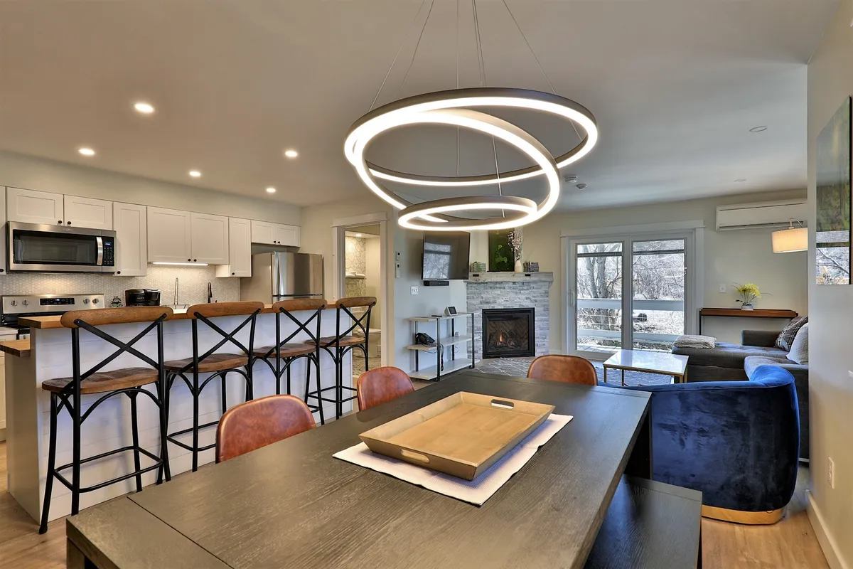 The image showcases a stylish, newly renovated condo interior featuring a gleaming kitchen island, a modern chandelier that adds a touch of elegance, a cozy fireplace perfect for relaxing after a day on the slopes, and a spacious dining table ideal for gatherings. The ambiance is warm, inviting, and contemporary, highlighting spaces suited for both entertaining and quiet moments.