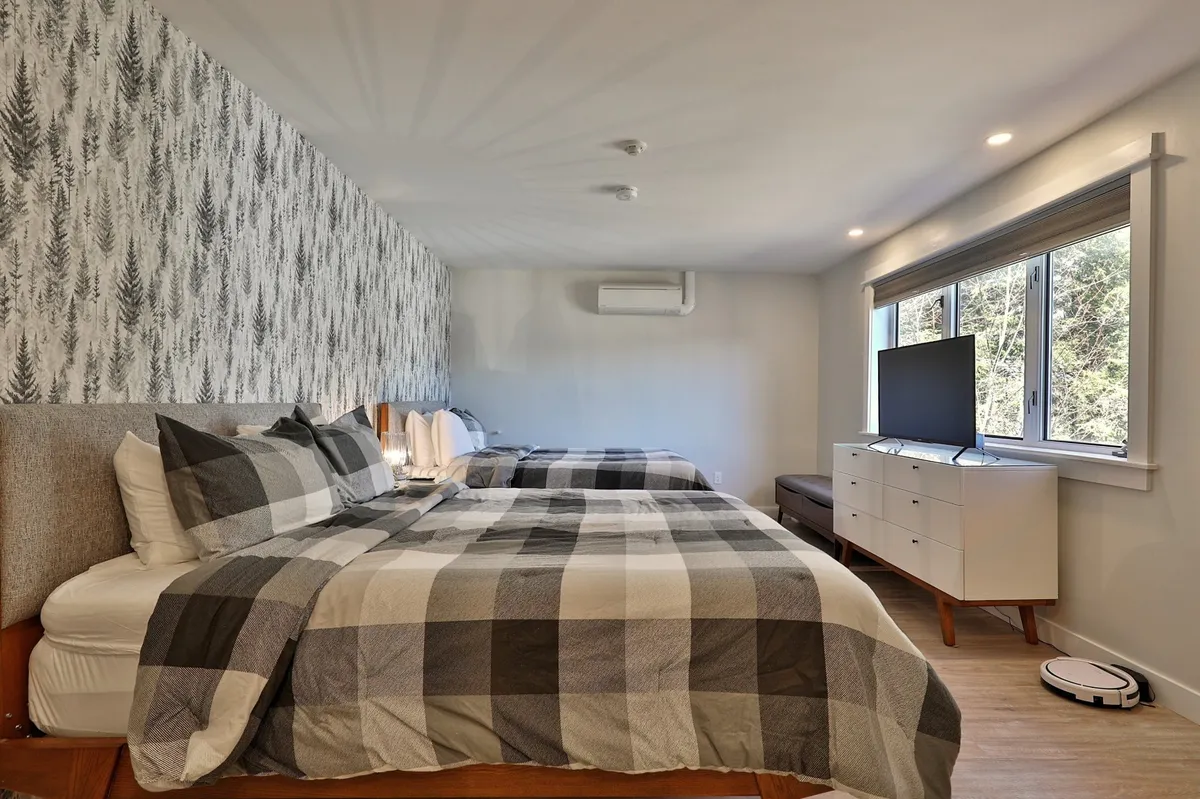 This image displays a spacious bedroom with two large beds dressed in plaid bedding, a decorative fern-patterned wallpaper, a large window letting in ample natural light, a flat-screen TV, and a minimalist dresser. The room exudes comfort and a connection to nature through its tranquil décor.