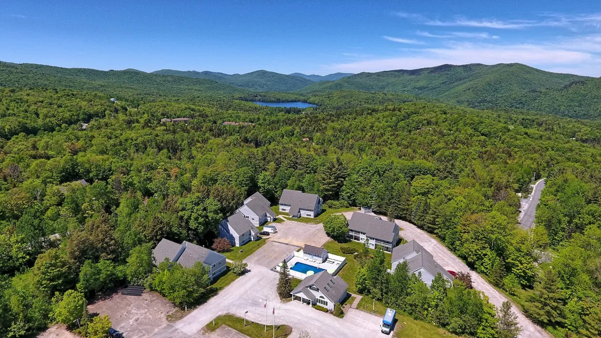An inviting exterior view of the residential complex, featuring a swimming pool, multiple buildings nestled in a forested landscape, and a scenic mountain backdrop. The setting conveys tranquility, privacy, and connection to nature, with hints of both summer and winter recreational activities.