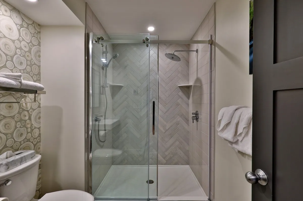 A modern bathroom featuring a glass shower enclosure with a distinctive herringbone tile pattern, a decorative wall with a wood slice design, and neatly arranged towels. The space feels serene and is clearly designed for self-care routines.