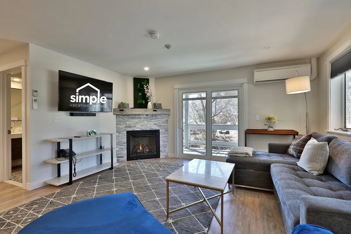 A sunlit living area featuring a stylish modern gas fireplace as the focal point, with a plush sofa, coffee table, and smart TV. Expansive windows frame a scenic outdoor view, enhancing the warm, inviting atmosphere. The overall design encourages relaxation and socializing.