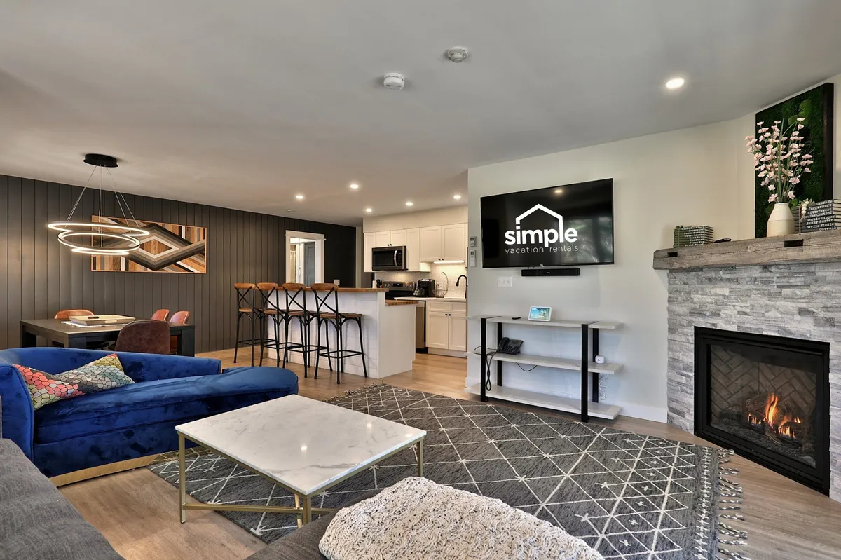 A cozy and modern living area with a plush blue velvet sofa, a fireplace, a dining area with bar stools, and a TV displaying 'simple vacation rentals.' The space invites relaxation and gatherings, evoking homelike comfort and the promise of restful, quiet evenings.