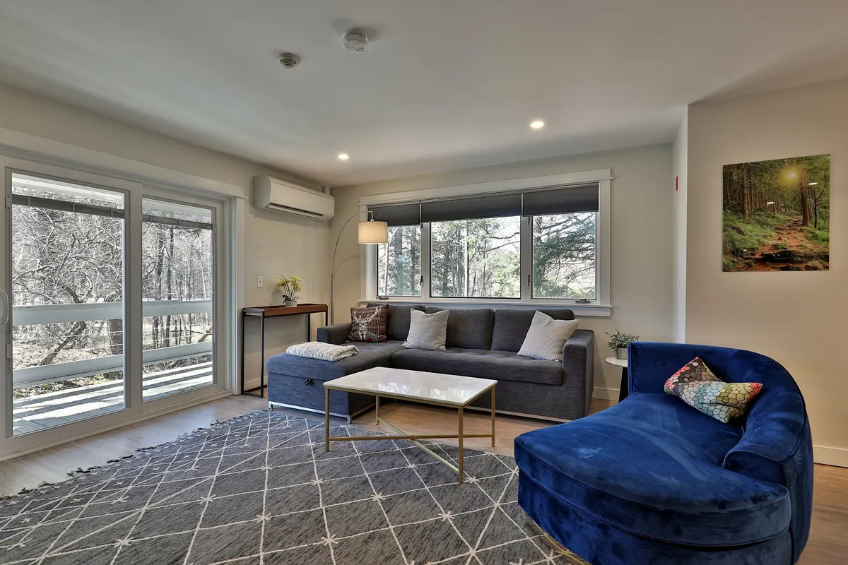 A cozy corner of the living area featuring a blue armchair, gray sectional sofa, and a coffee table. Large windows flood the room with natural light and offer serene views of the outdoors, while tasteful wall art adds to the inviting atmosphere.