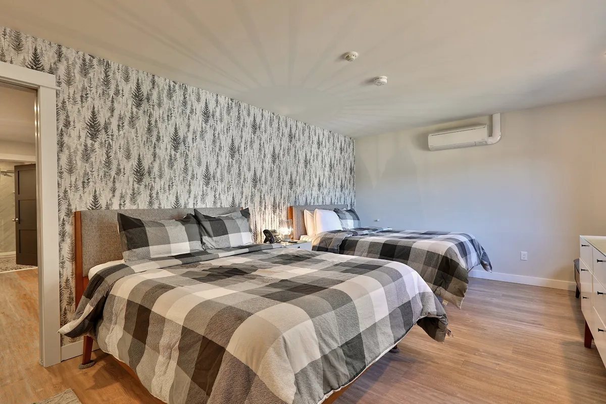 This image captures a cozy bedroom with plaid bedding, fern-patterned wallpaper, and soft bedside lighting. The mood is warm and inviting, suggesting a peaceful retreat after a day exploring Vermont's beauty.
