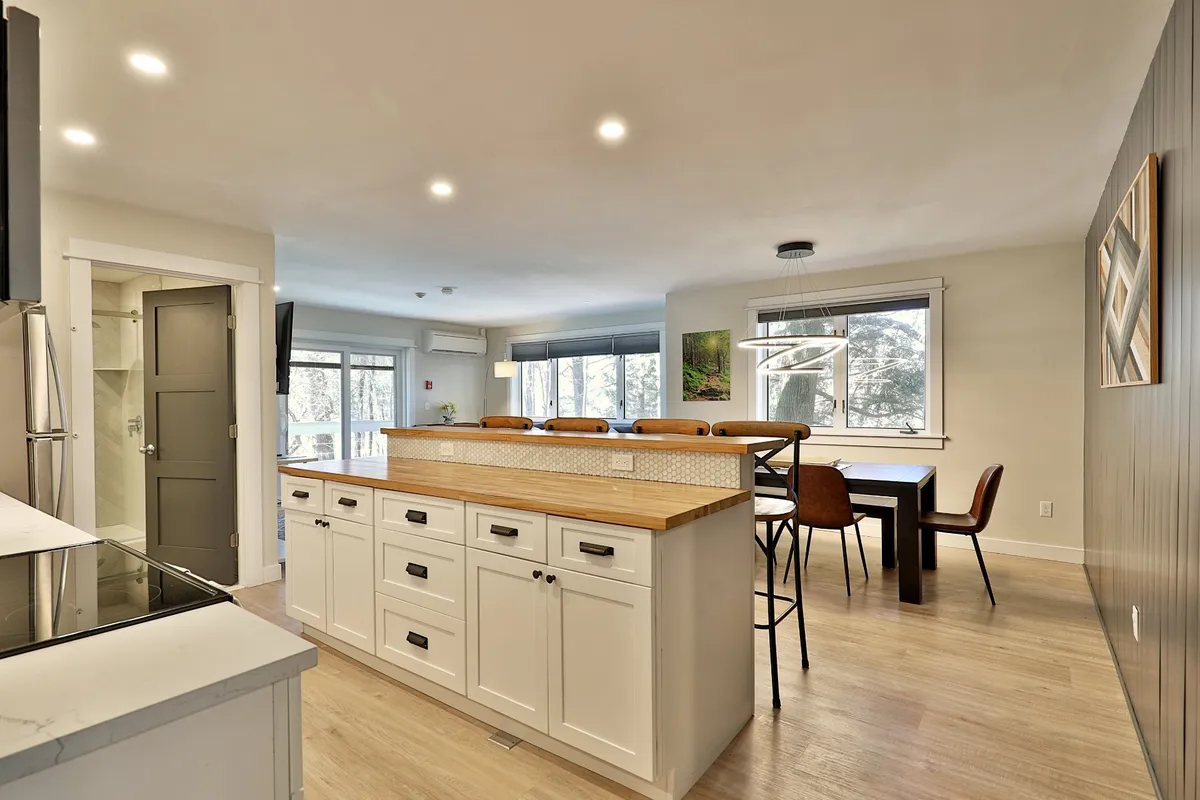 A wide-angle view of the kitchen and dining area, featuring a kitchen island and a connected dining space with large windows for natural light. The setup is designed for cooking, family gatherings, and socializing over meals, exemplifying openness and connectivity.