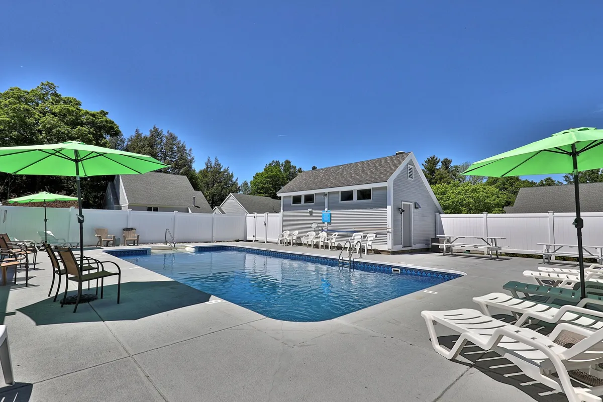 This exterior image highlights the outdoor swimming pool surrounded by lounge chairs, green umbrellas, and a pool house, set against a background of leafy trees. The area is designed for sunbathing, relaxing, and socializing, with ample space for family gatherings and poolside barbecues.