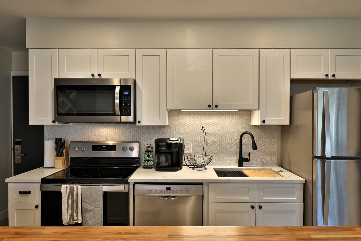 A bright, contemporary kitchen featuring stainless steel appliances, white cabinetry, a wooden countertop, and a black sink. Morning sunlight streams in, highlighting a welcoming space ideal for starting the day with coffee before heading out.