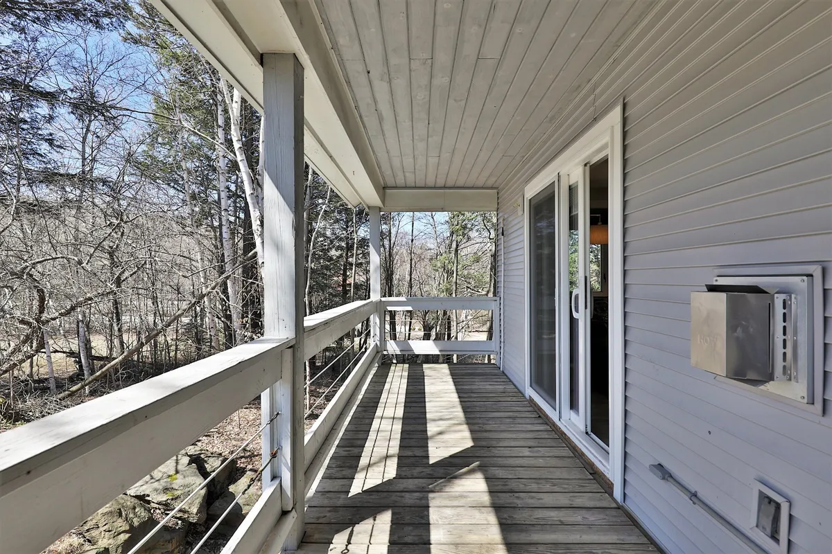 A private balcony framed by a wooden railing and surrounded by lush trees, offering a peaceful outdoor retreat just off the main living space. The deck flooring and natural setting provide an ideal spot for fresh air and relaxation.